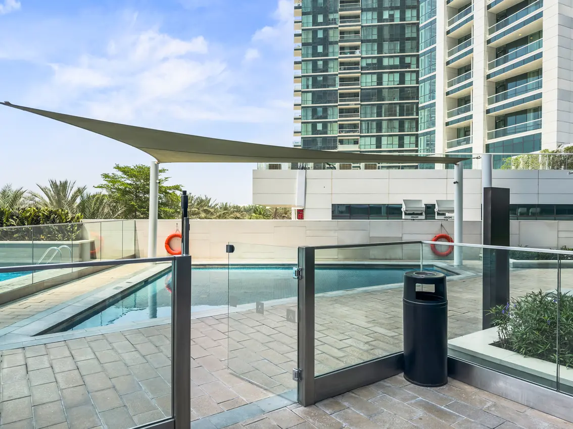 Rooftop pool area with glass railings, a tan shade sail, and a high-rise building in the background.