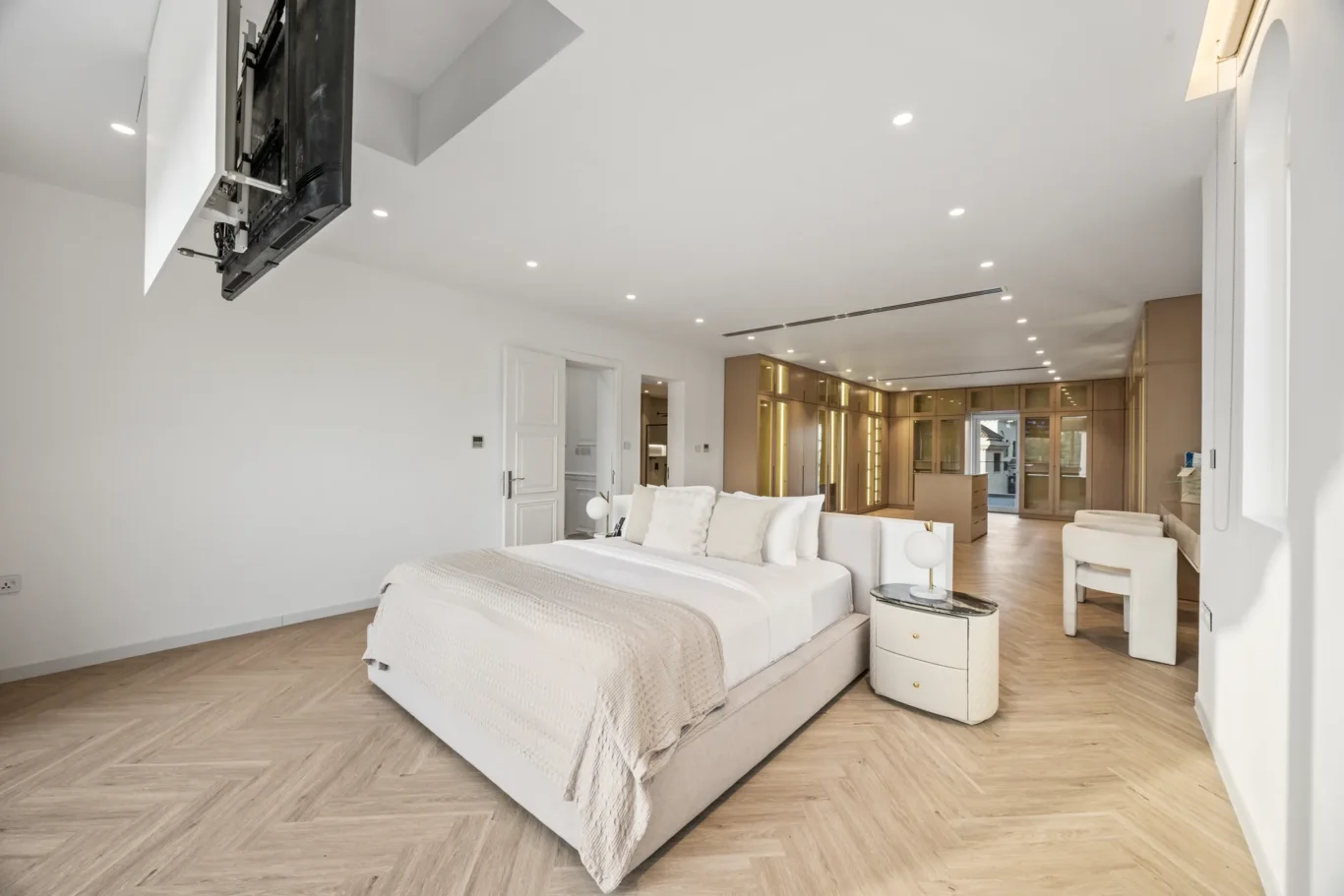 Luxurious white bedroom with a large bed and beige bedding; a ceiling-mounted TV extends from the wall at the foot of the bed.