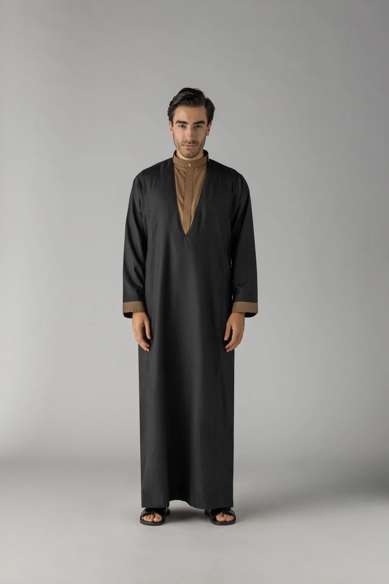 Man standing in a long black abaya with a brown inner collar and cuffs, against a neutral gray background.