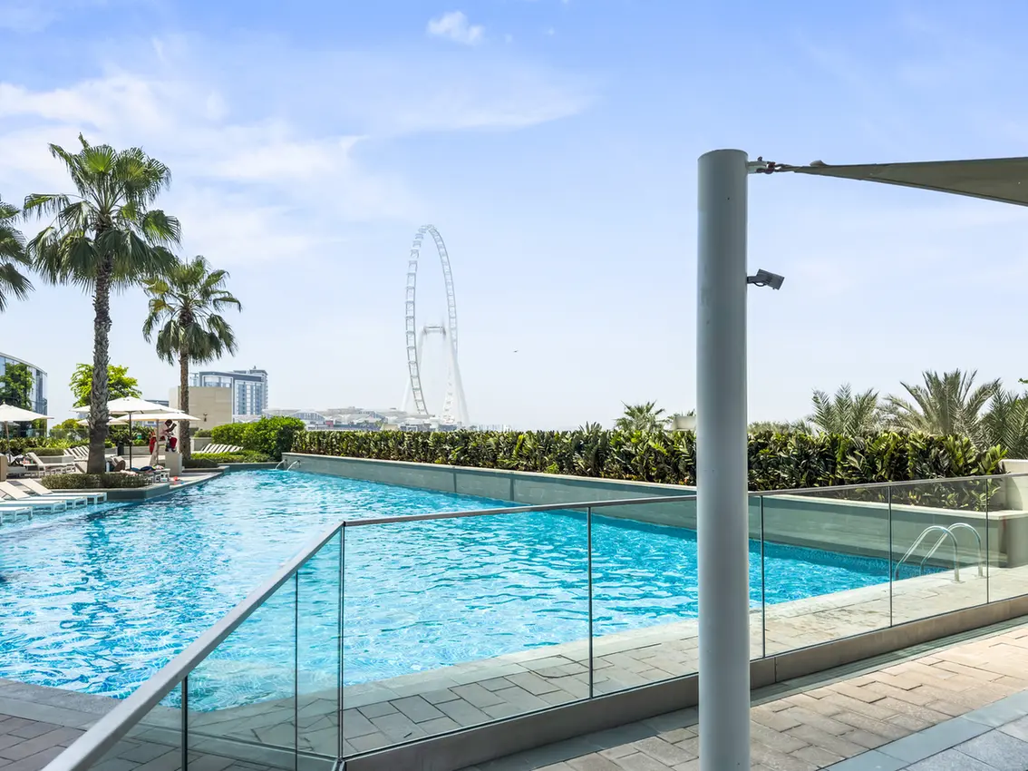 Outdoor pool area with glass railing, palm trees, and a clear blue sky; a large Ferris wheel visible in the distance.