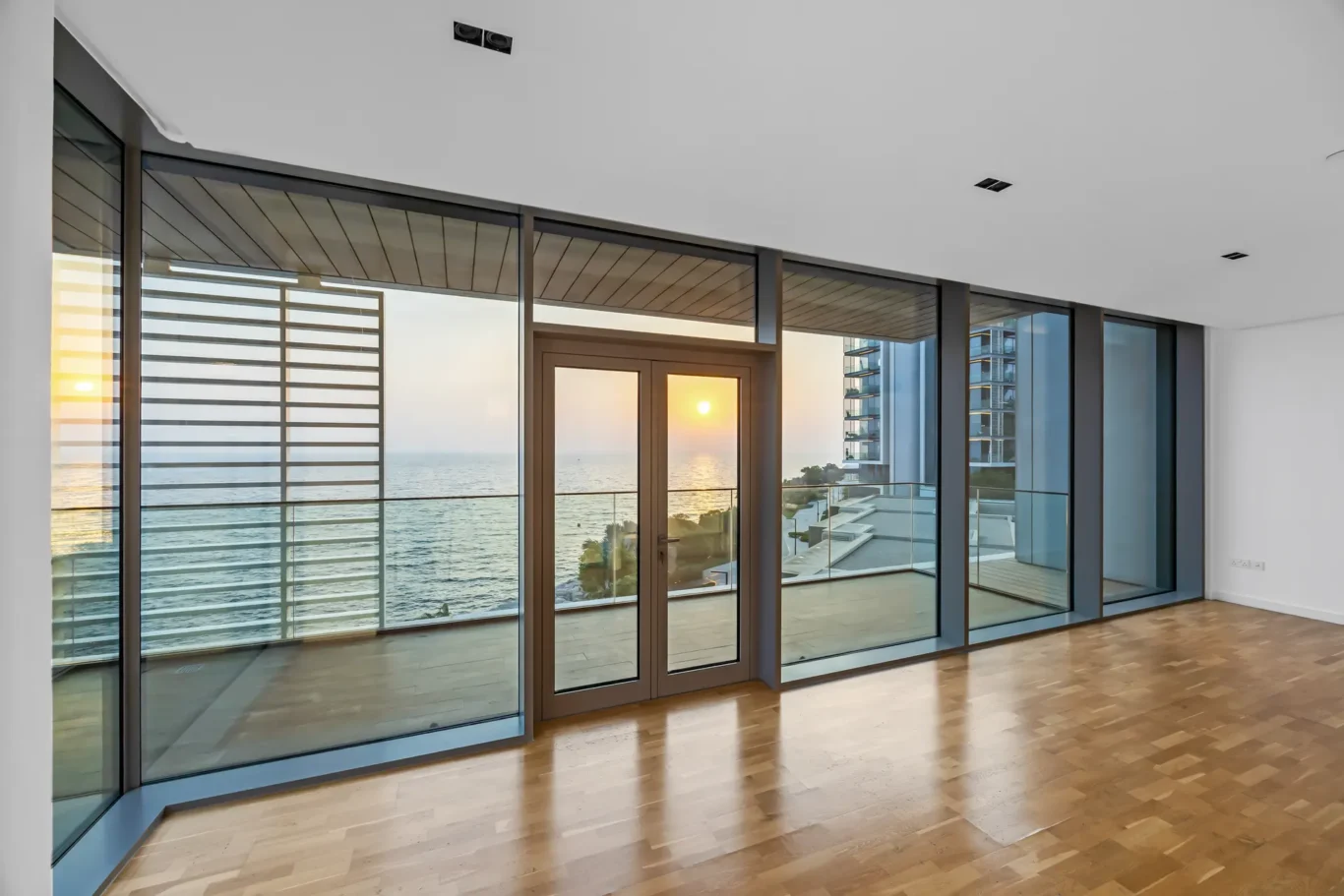 Interior of a bright modern room with large glass doors overlooking the ocean at sunset; wood floor and minimal decor.