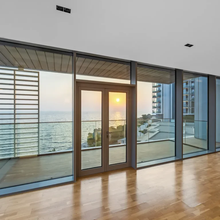 Interior of a bright modern room with large glass doors overlooking the ocean at sunset; wood floor and minimal decor.