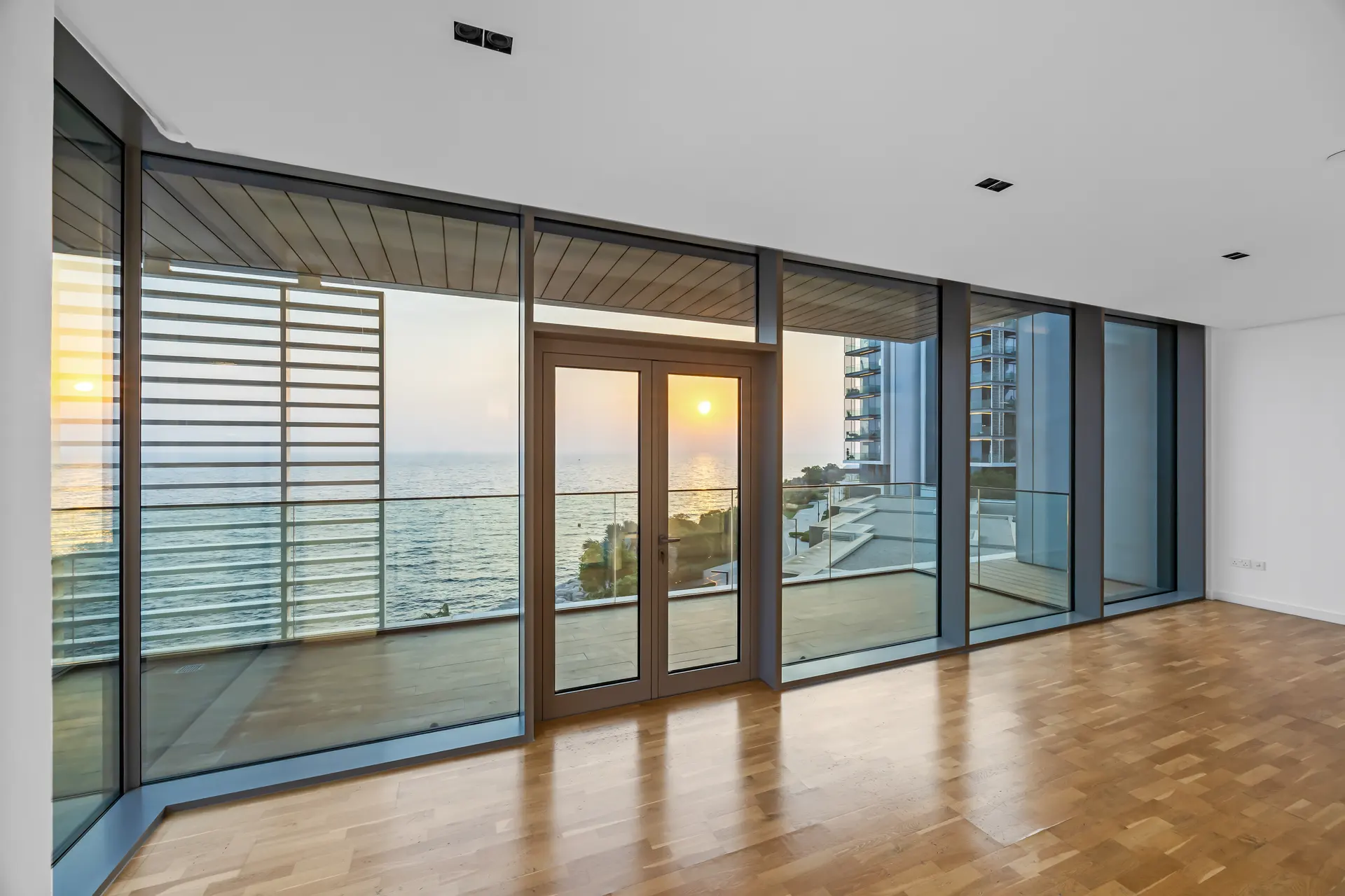 Interior of a bright modern room with large glass doors overlooking the ocean at sunset; wood floor and minimal decor.