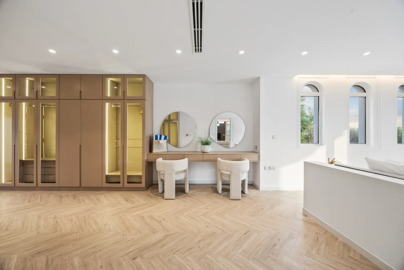 Modern dressing area with tan wardrobes, round mirrors above a beige vanity, two upholstered stools, and large arched windows.