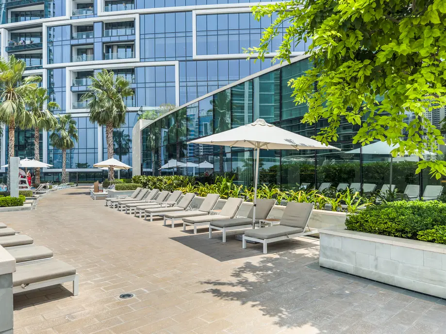 Modern hotel poolside terrace with beige lounge chairs, white parasols, and a glass-front building in the background.
