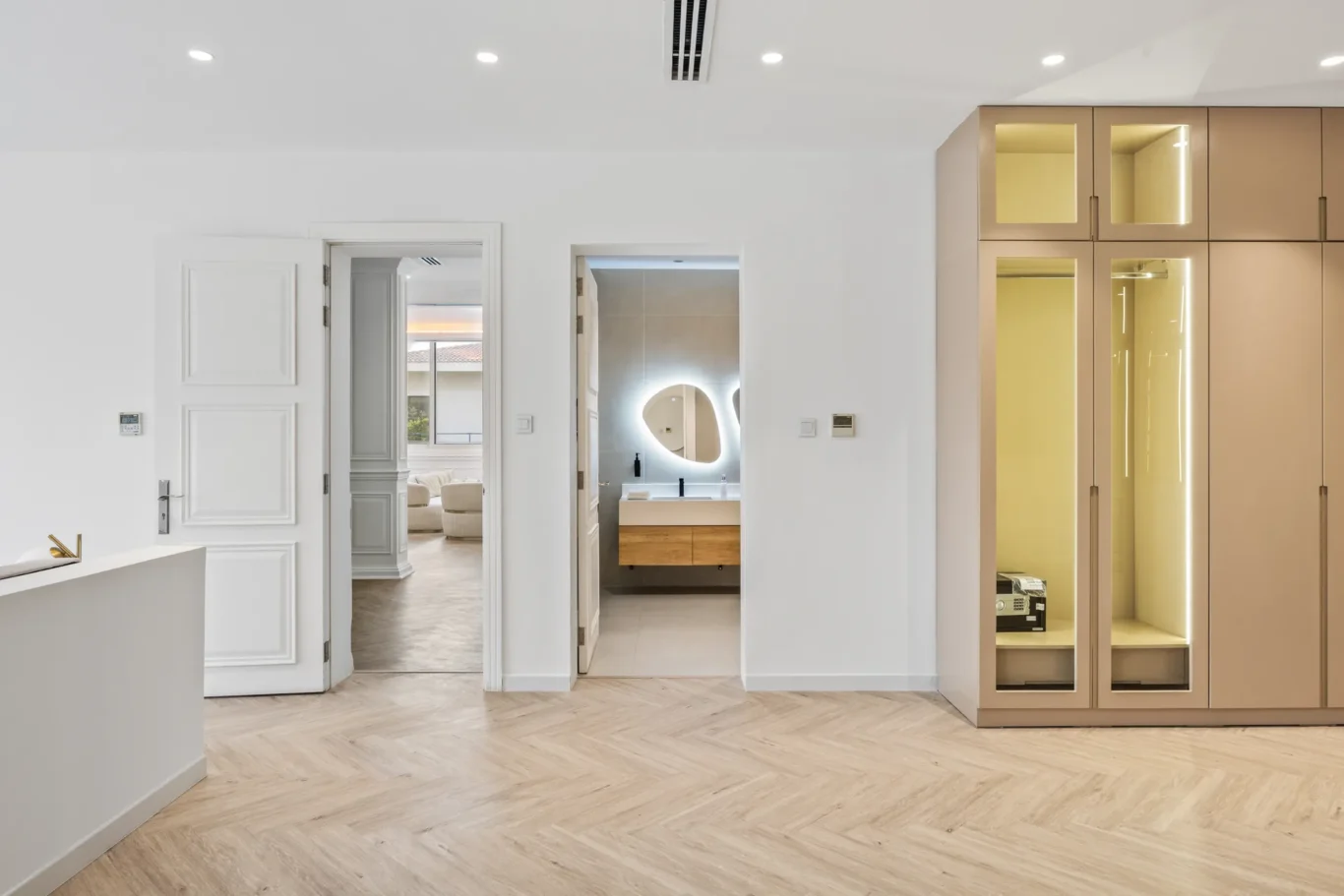 Modern white interior featuring two open doorways: a bathroom with a backlit mirror and wooden vanity on the right, and beige floor-to-ceiling storage cabinets to the far right.