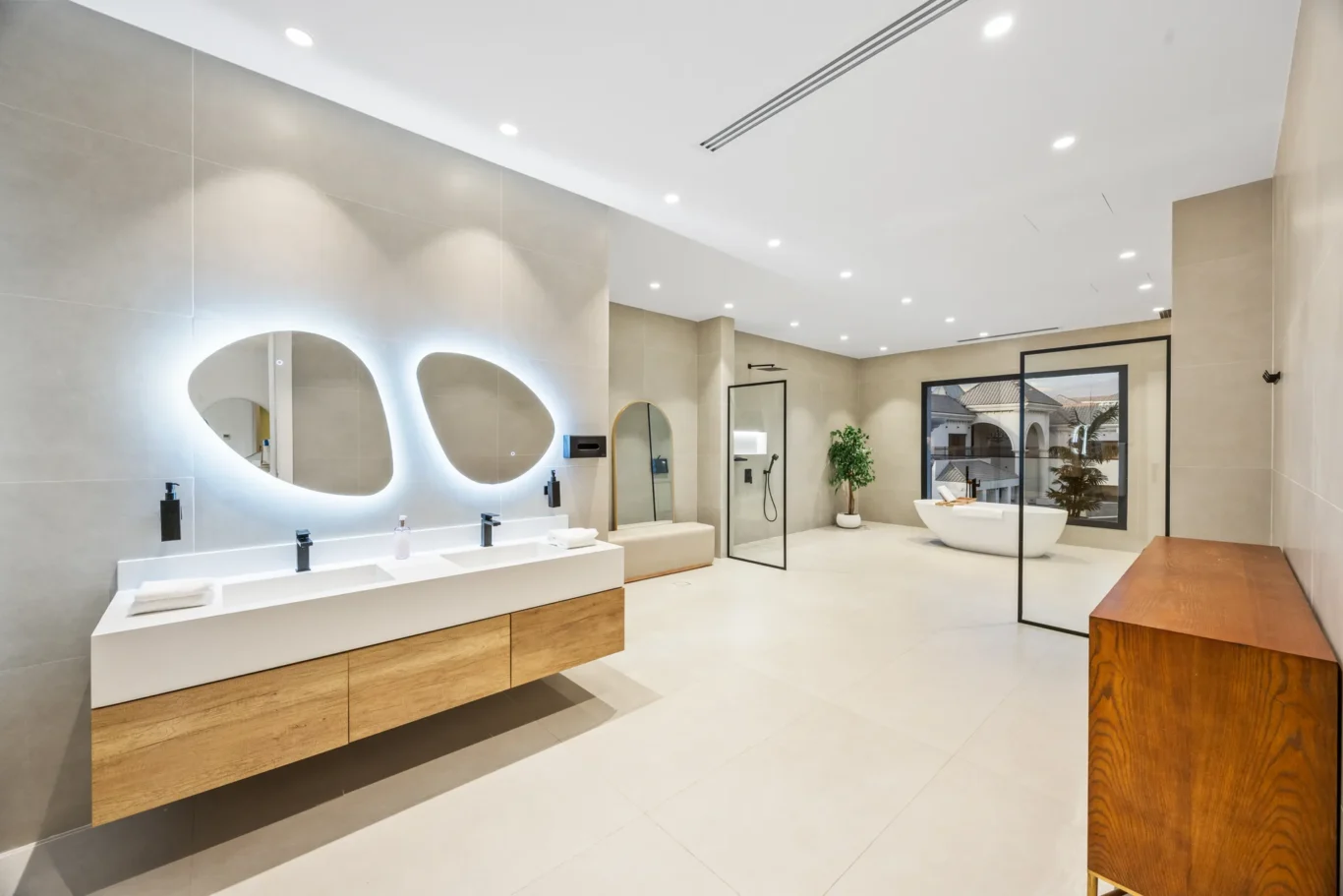 Modern spa-style bathroom with a double white vanity, wood drawer bases, and backlit oval mirrors.