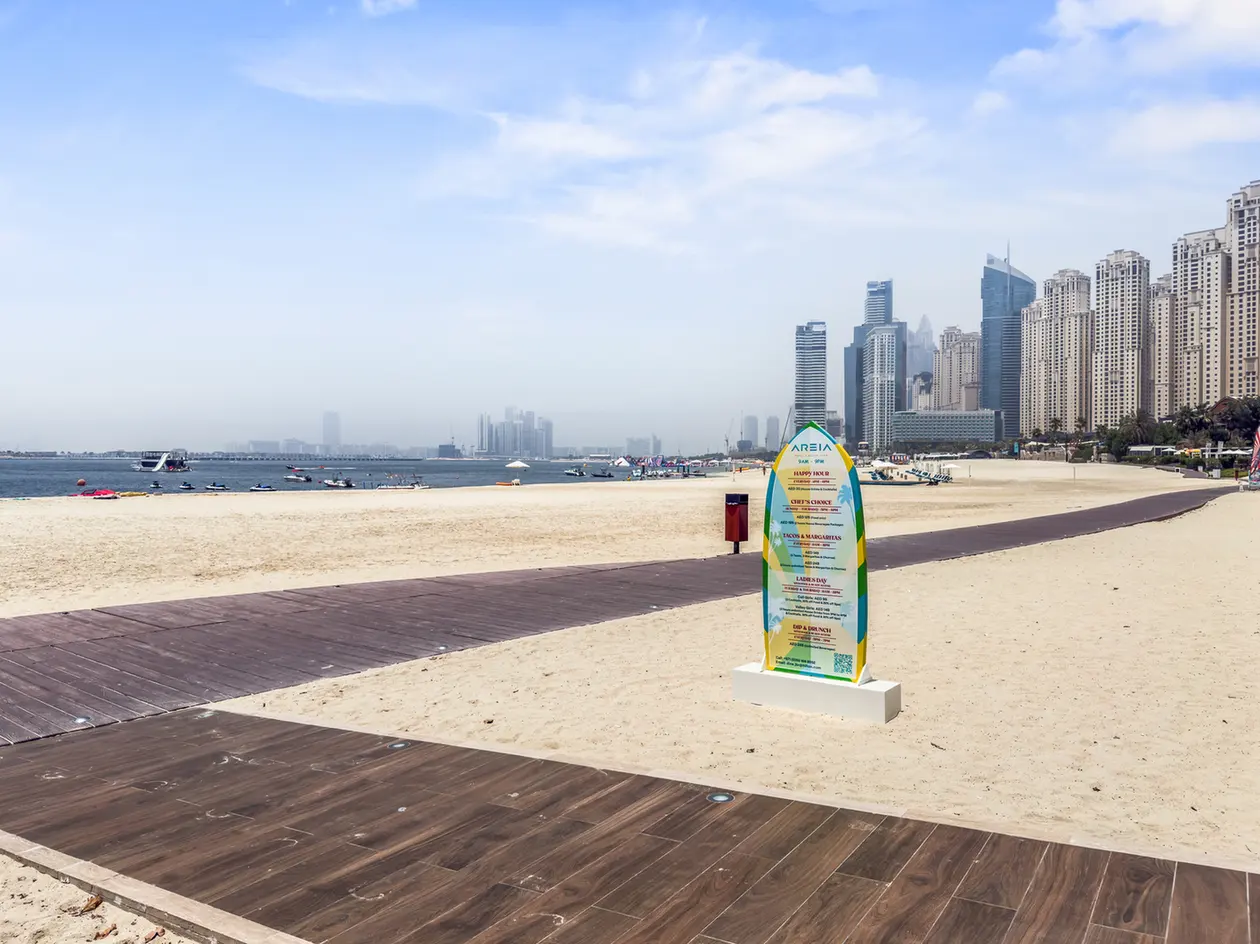 Sunny beach with a wooden boardwalk, calm water, and a colorful freestanding sign in the foreground; tall city skyscrapers line the coast in the distance.
