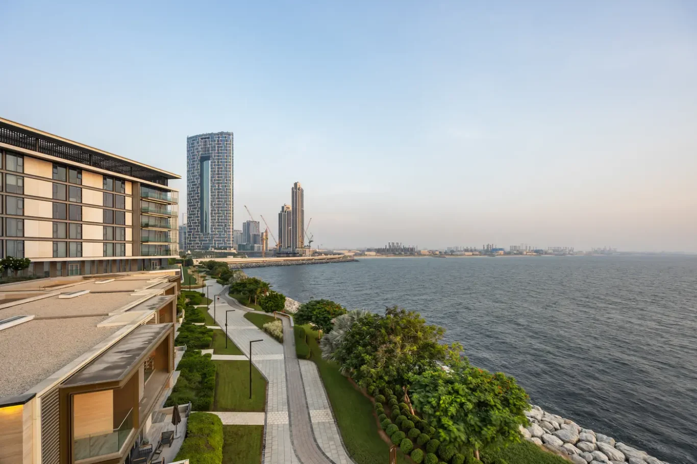 Modern waterfront promenade with landscaped gardens and a glassy bay, high-rise buildings in the background under a clear sky