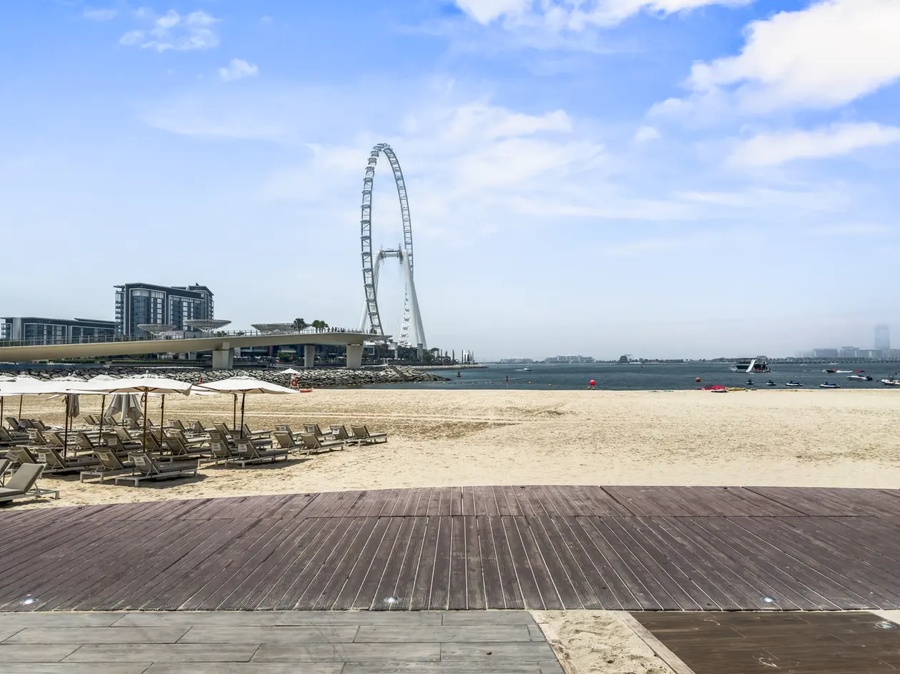 Beach scene with rows of loungers under umbrellas along the shore, a wooden boardwalk in the foreground, and a large circular observation wheel in the bay (Ain Dubai).