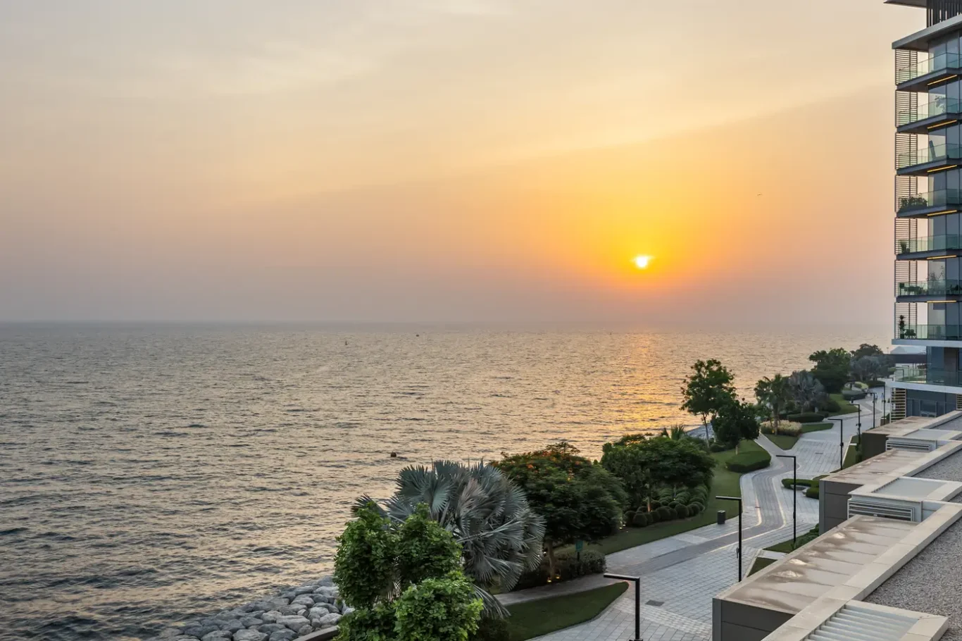 Sunset over the calm sea seen from a coastal promenade beside a modern glass building on the right.