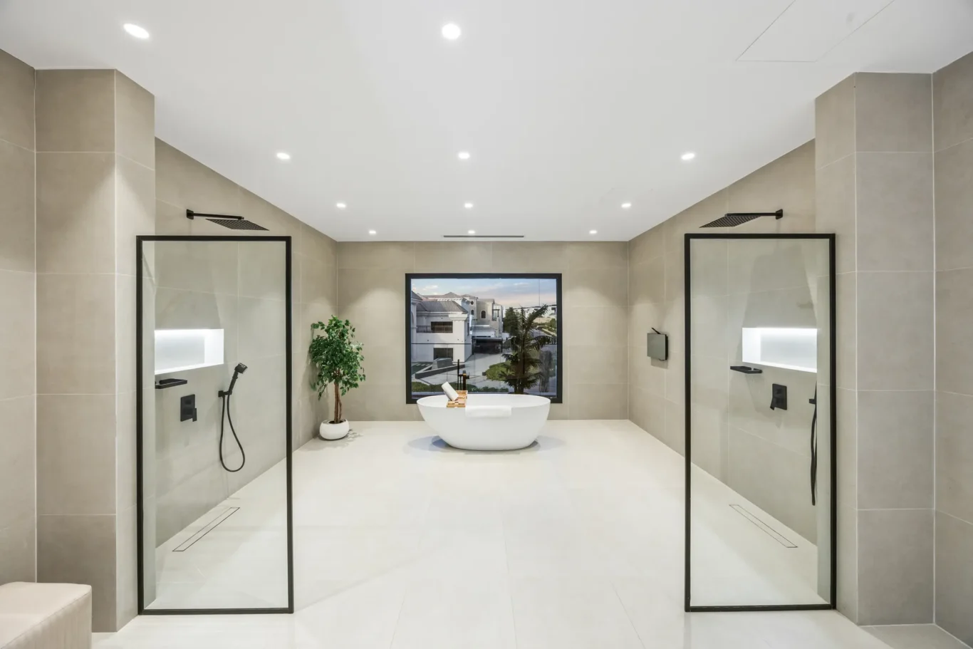Symmetrical bathroom with two glass-enclosed showers flanking a white freestanding tub and a large window overlooking outdoor buildings.
