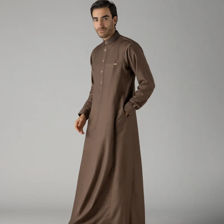 Man wearing a brown ankle-length thobe with black sandals stands against a gray backdrop, mid-stride to the left side of the frame.