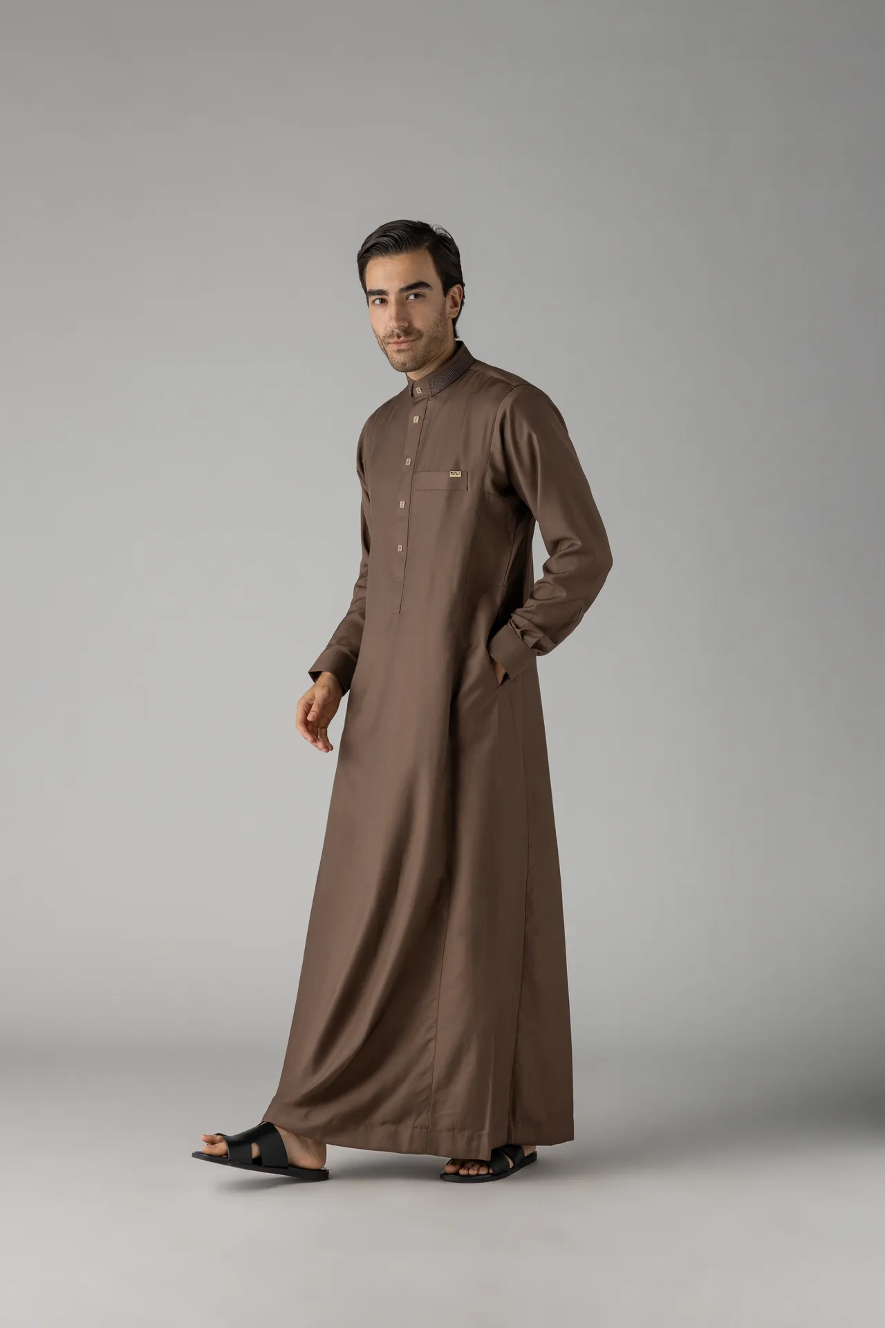 Man wearing a brown ankle-length thobe with black sandals stands against a gray backdrop, mid-stride to the left side of the frame.