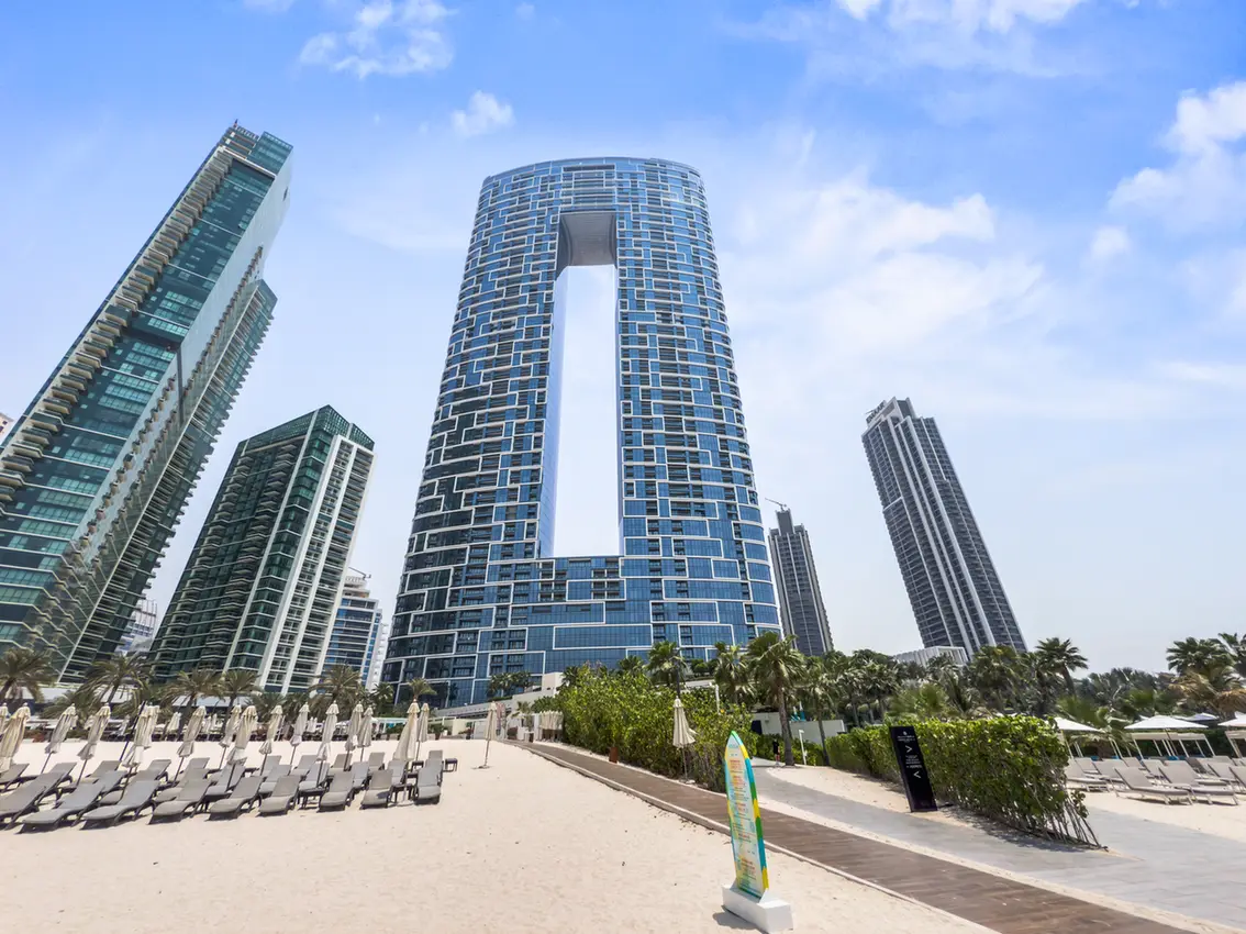 Curved glass skyscraper with a large central opening, flanked by other tall blue towers on a sunny beach scene with lounge chairs and palm trees nearby.] ,
