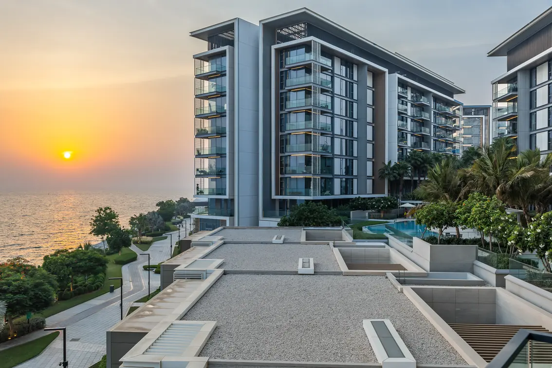 Modern waterfront residential towers at sunset with glass balconies and palm trees by the sea.