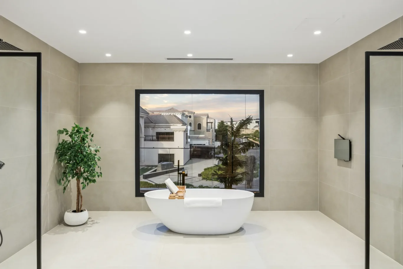Modern bathroom with freestanding white tub, large window, beige tile walls, and a potted plant.