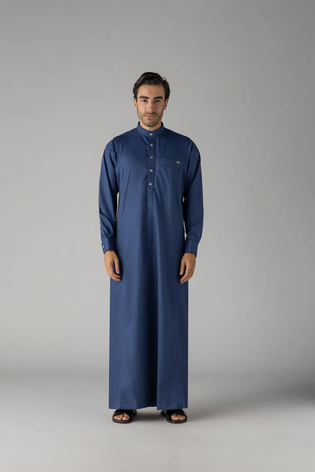 Man standing in a blue thobe against a gray studio background.