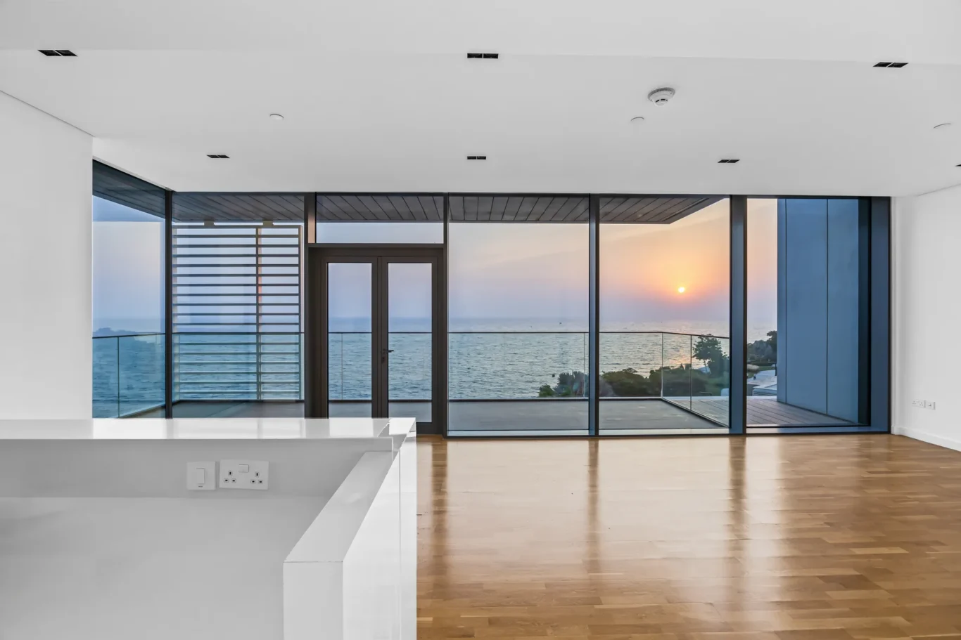 Open-plan living area with large glass doors opening to a sunset over the sea, white surfaces, and light wooden floors.