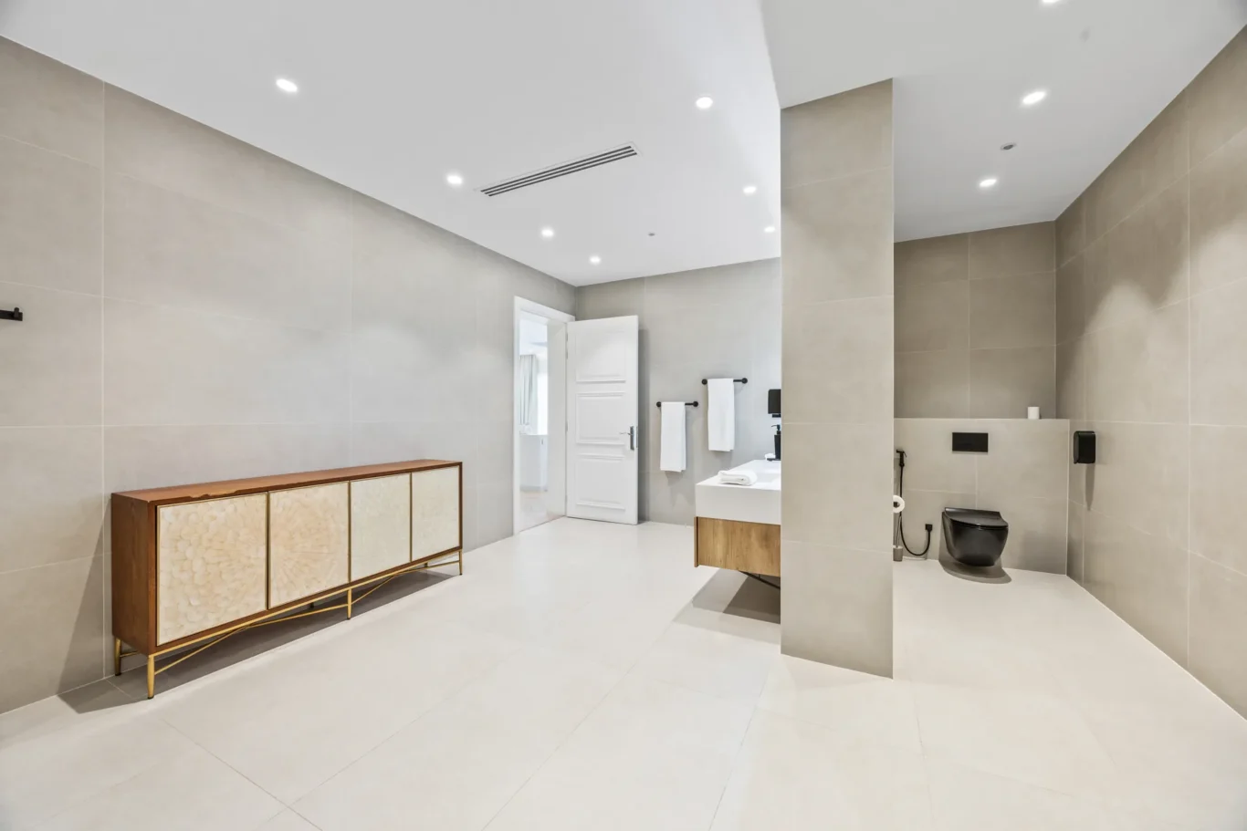 Spacious modern bathroom with beige tiled walls, white fixtures, and a wooden cabinet along the left wall under recessed ceiling lights.