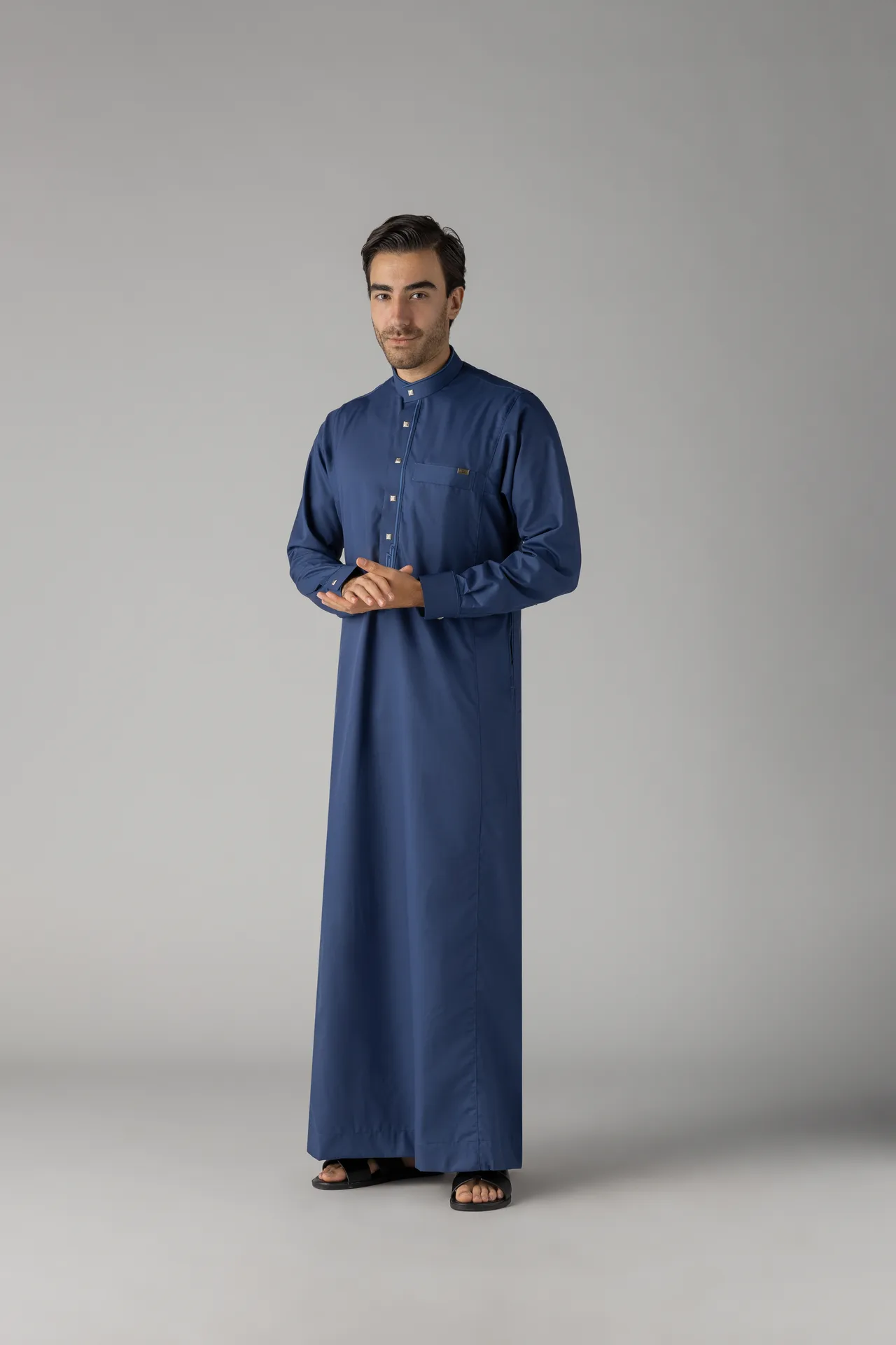 Full-body portrait of a man in a blue traditional thobe and black sandals, standing with hands clasped against a neutral grey backdrop