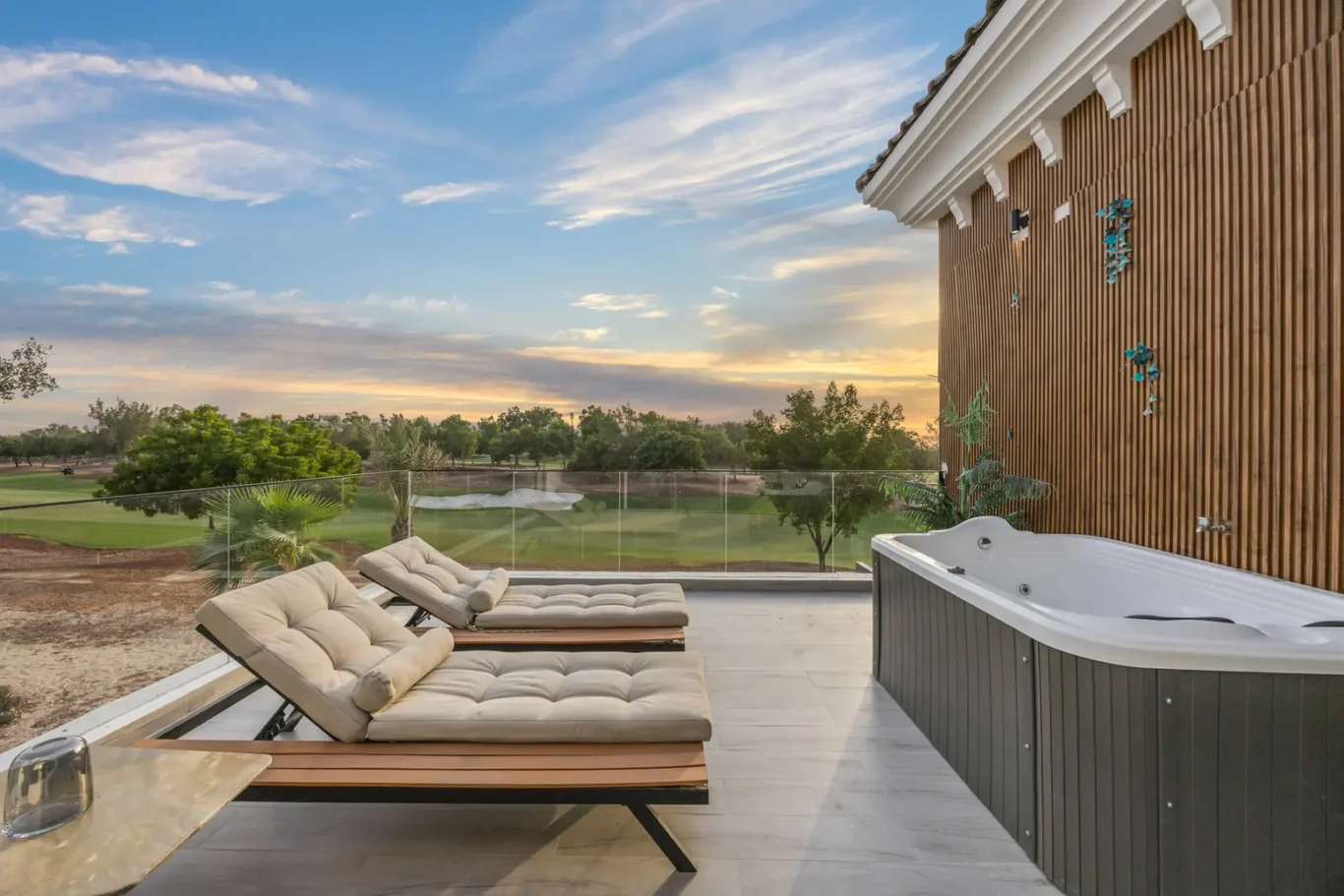 Outdoor patio with two cushioned lounge chairs and a hot tub, overlooking a green lawn at sunset.
