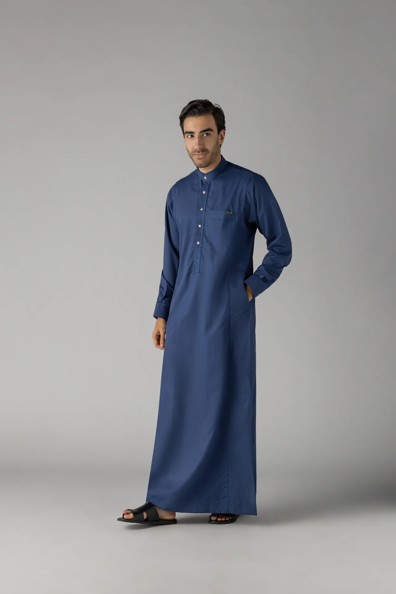 Man wearing a long blue thobe with a chest pocket and sandals, standing against a neutral gray backdrop.