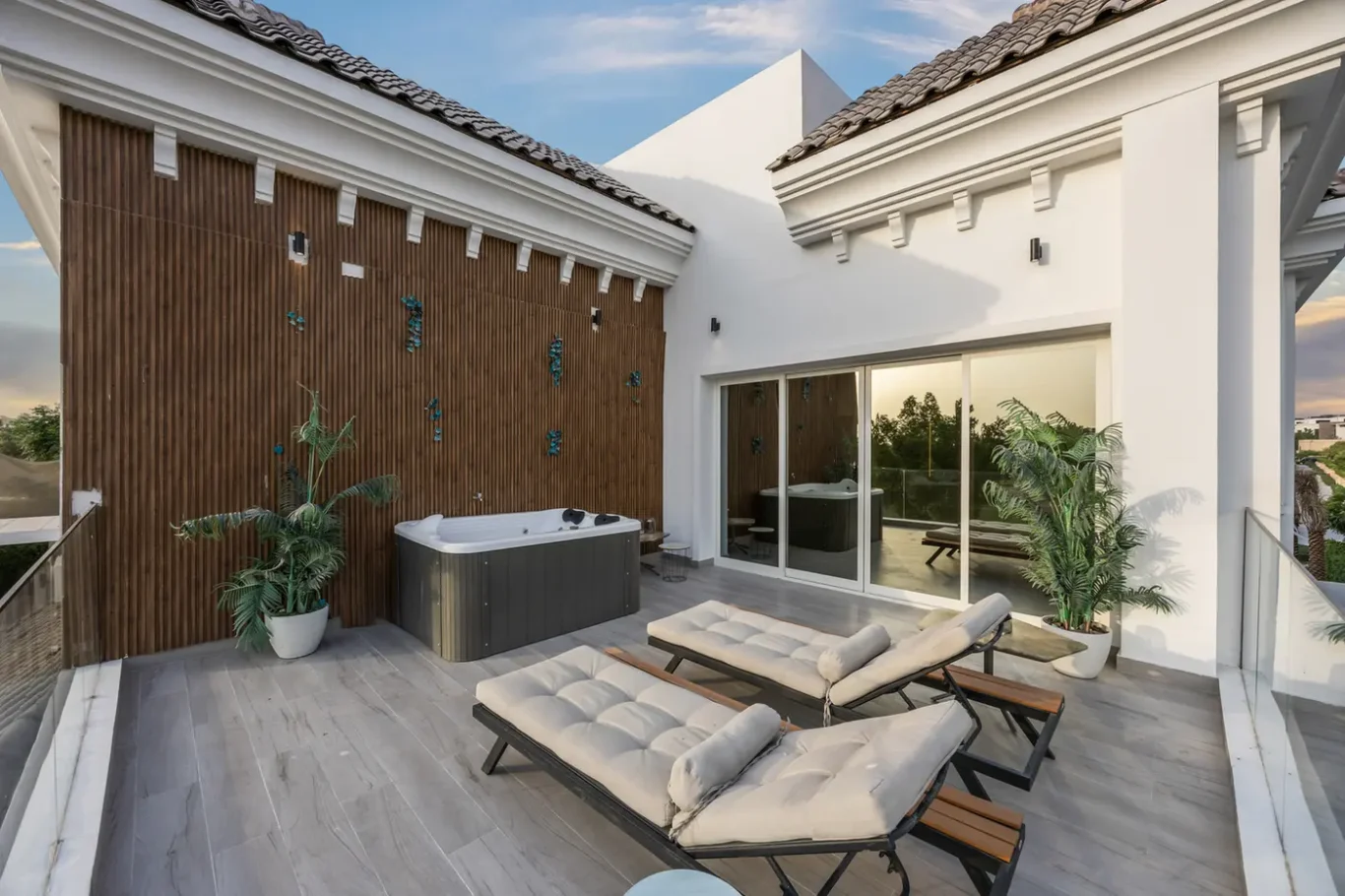Rooftop patio with a hot tub, three cushioned lounge chairs, a wooden slat privacy wall, and potted plants by sliding glass doors.