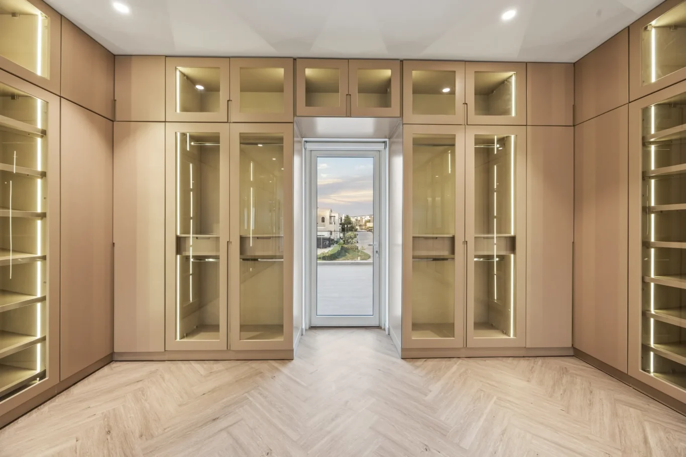 Walk-in wine cellar with beige glass-front cabinets, backlit shelves, and a door at the end showing an outdoor view.