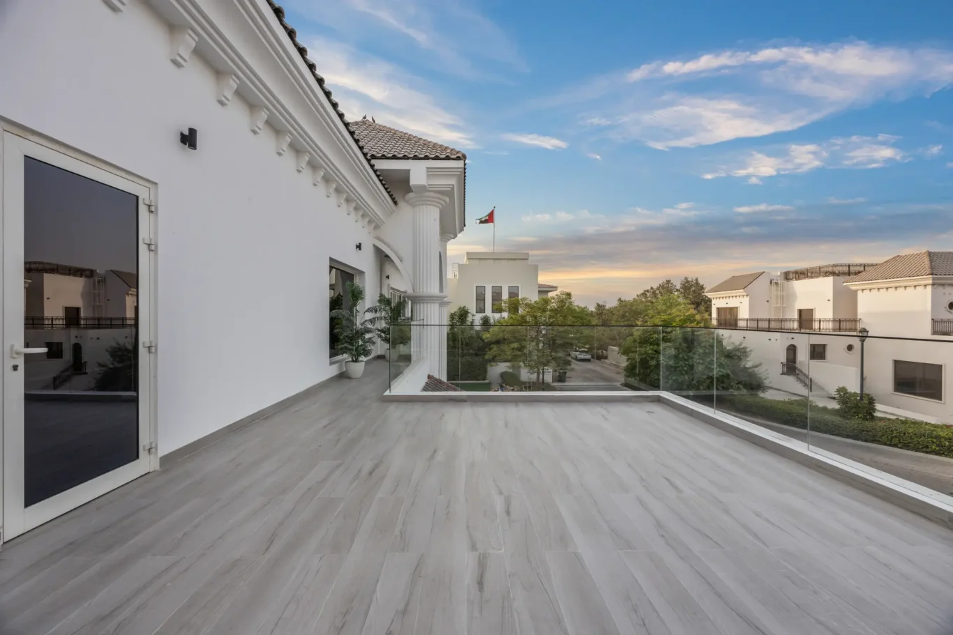 Spacious rooftop terrace with gray tile flooring, white walls, decorative molding, and a glass railing overlooking a leafy street. A distant Italian flag flies atop a neighboring building under a blue sky.