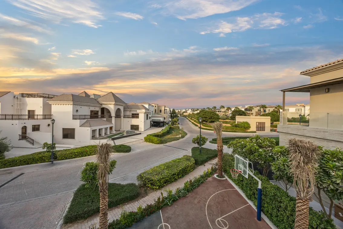 Sunset over a Mediterranean-style residential neighborhood with white villas, palm trees, and a small basketball court in the foreground.