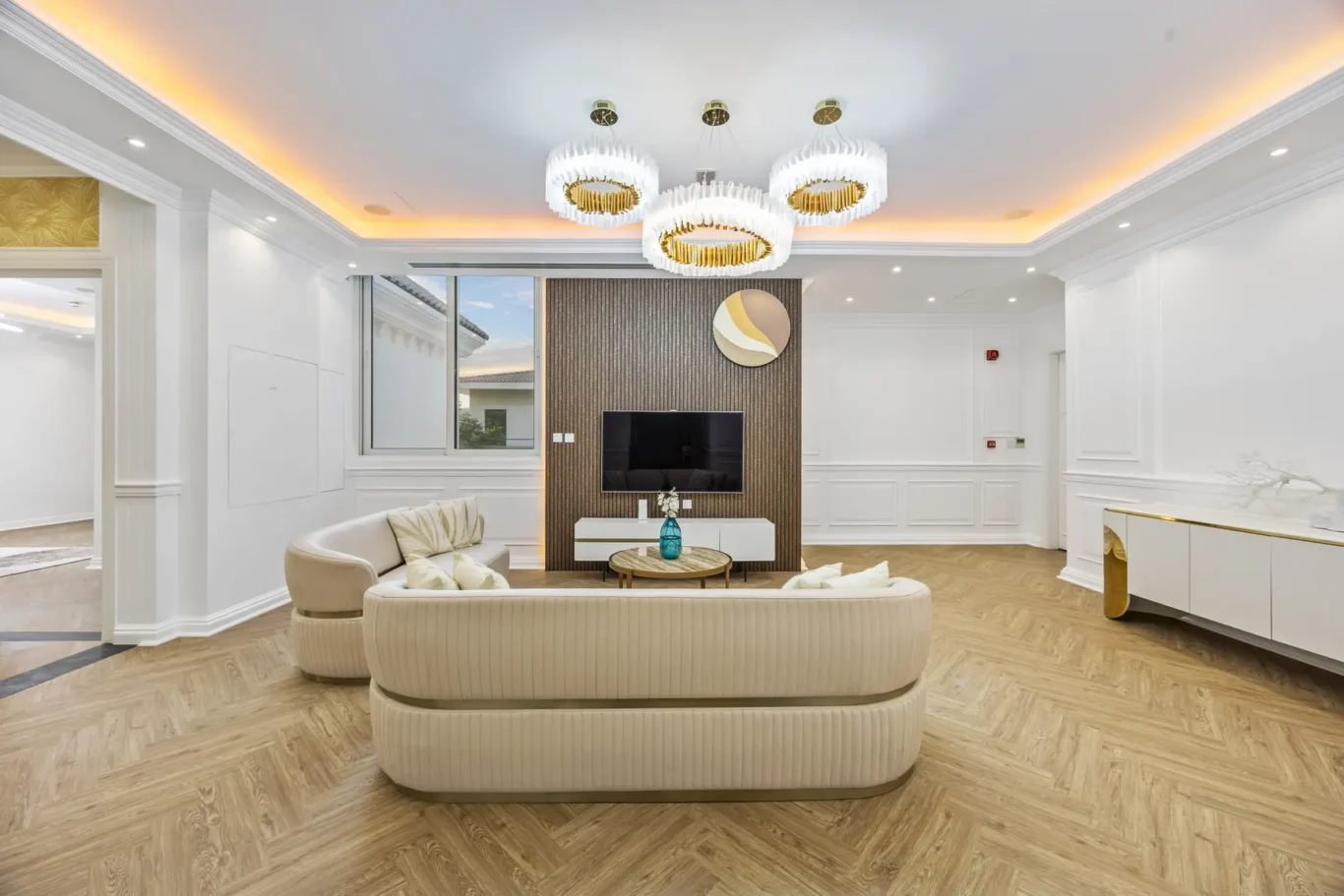 Modern living room with curved beige sofas, round coffee table, TV on dark wood panel, and a gold-ring chandelier against white walls.