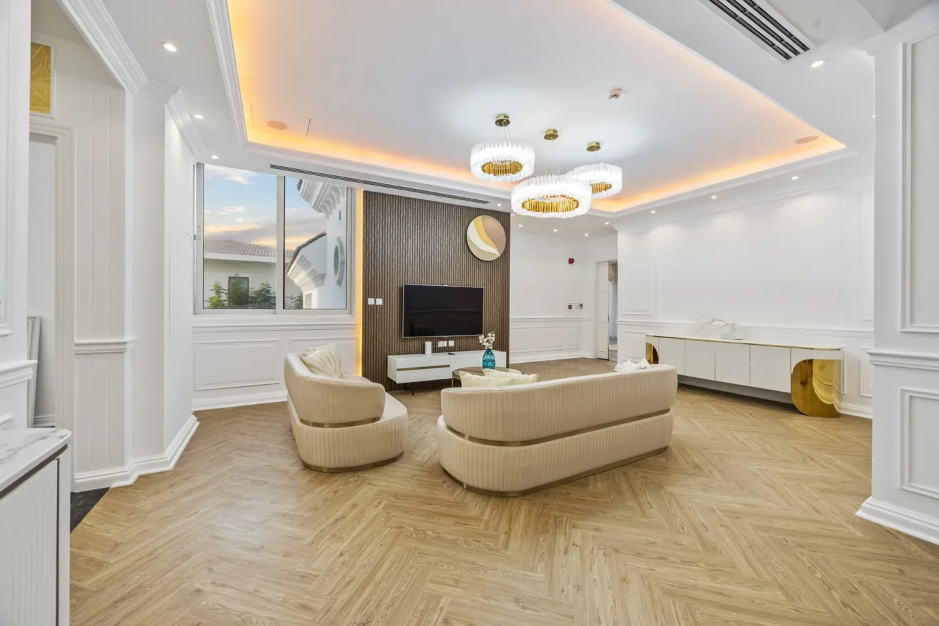 Bright modern living room with white walls, herringbone wood floor, curved beige sofas, wall-mounted TV, and circular pendant lights in gold rings.