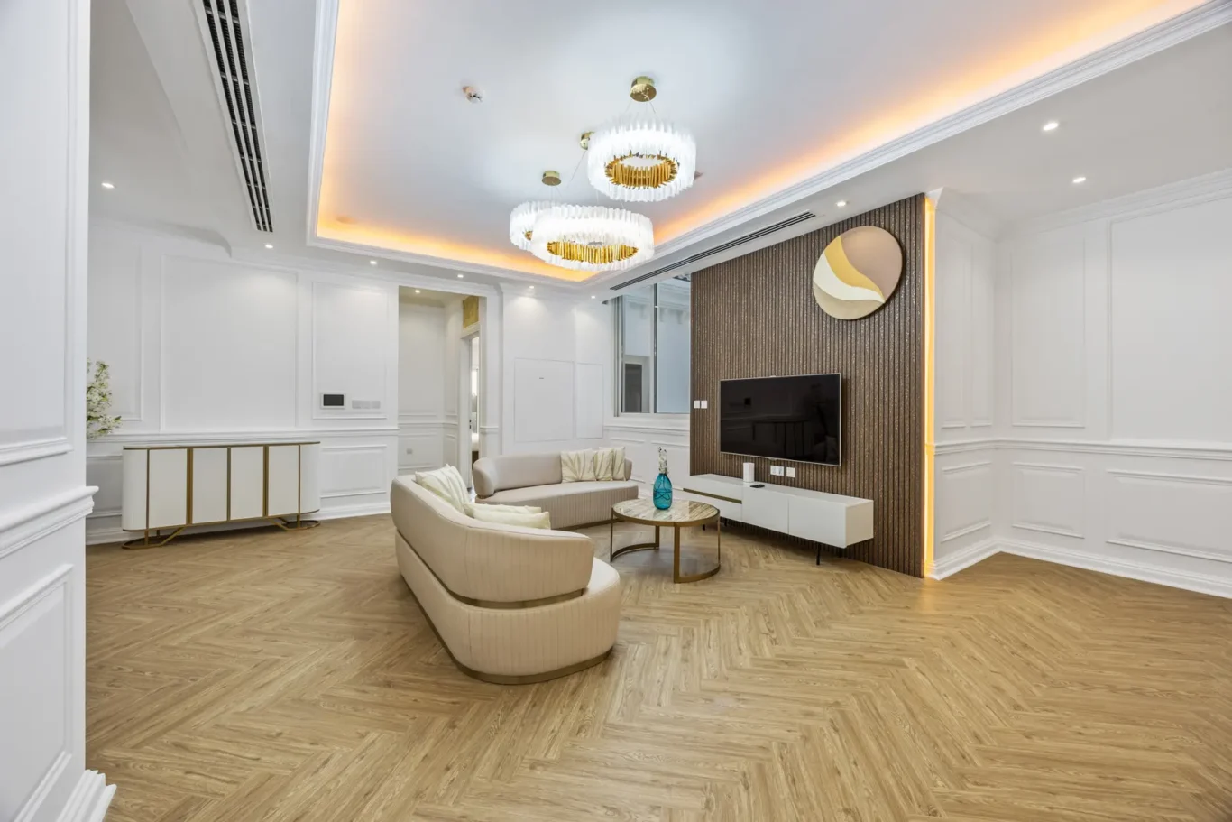 Elegant living room with white paneled walls, herringbone wood floor, cream curved sofas, and a dark textured accent wall behind a mounted TV.
