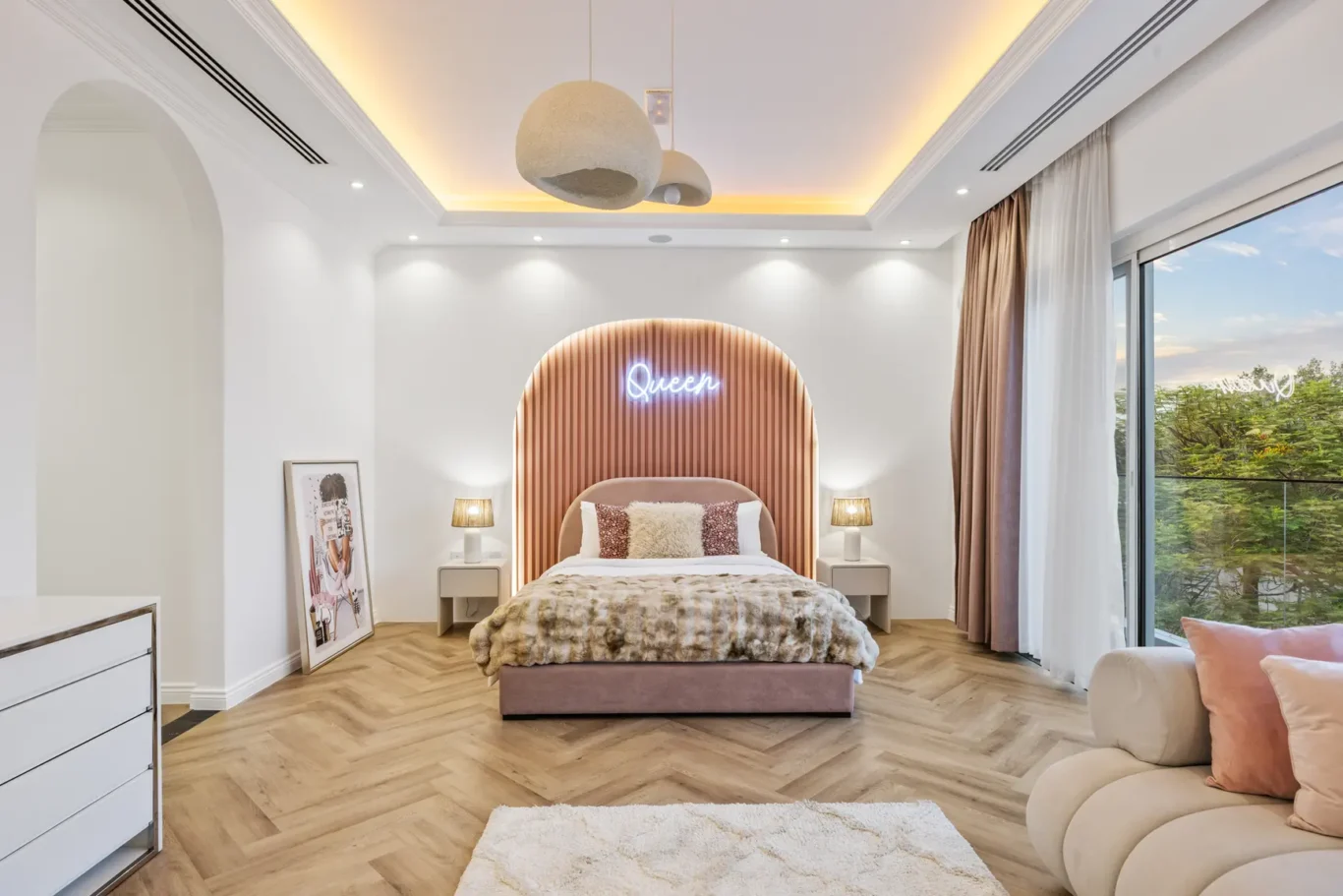Luxurious bedroom with a curved wood headboard, neon 'Queen' sign, two bedside lamps, and a plush bed in soft pink tones.