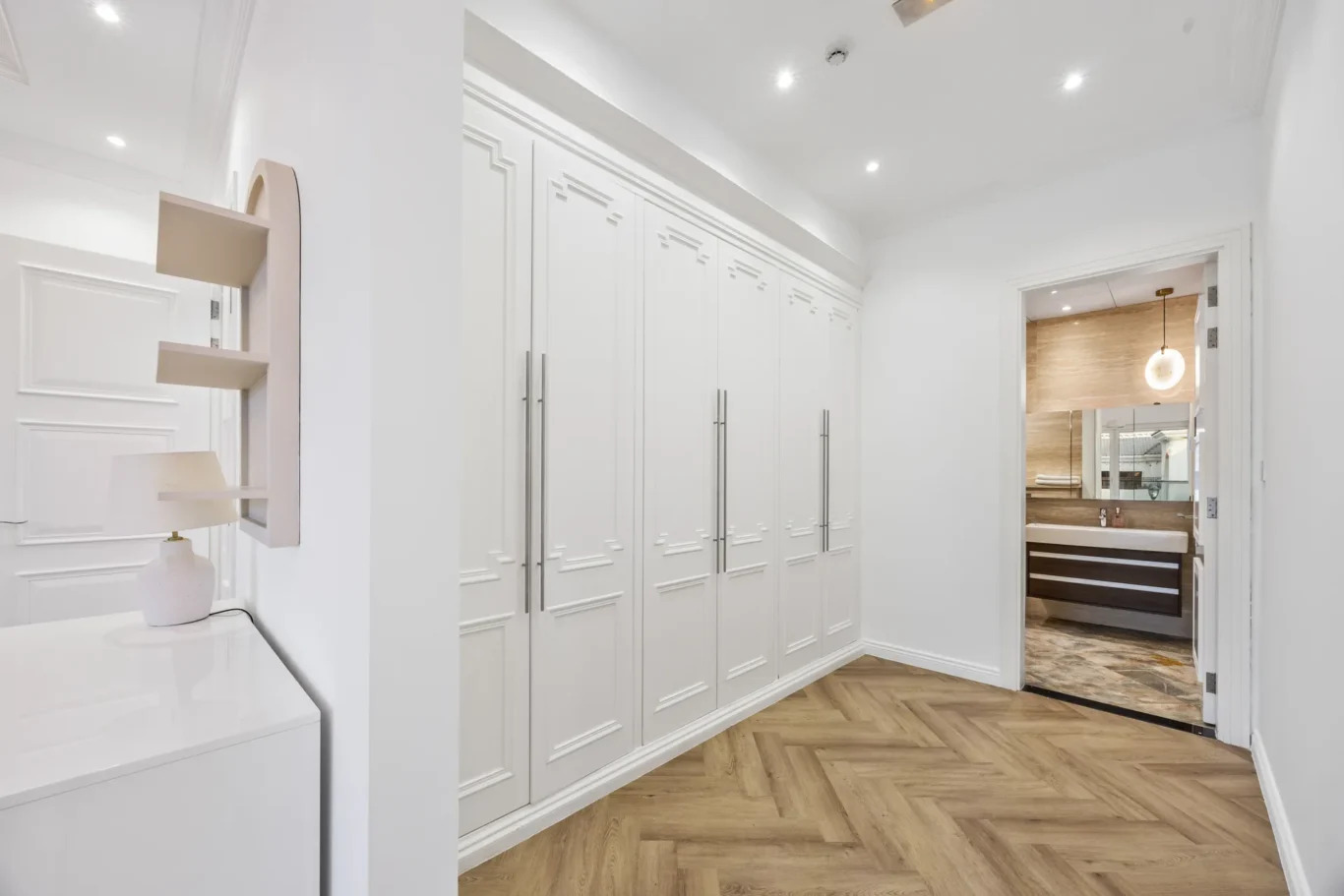 Bright hallway with tall white built-in wardrobes and long metal handles, herringbone wood floor, leading to a bathroom on the right.