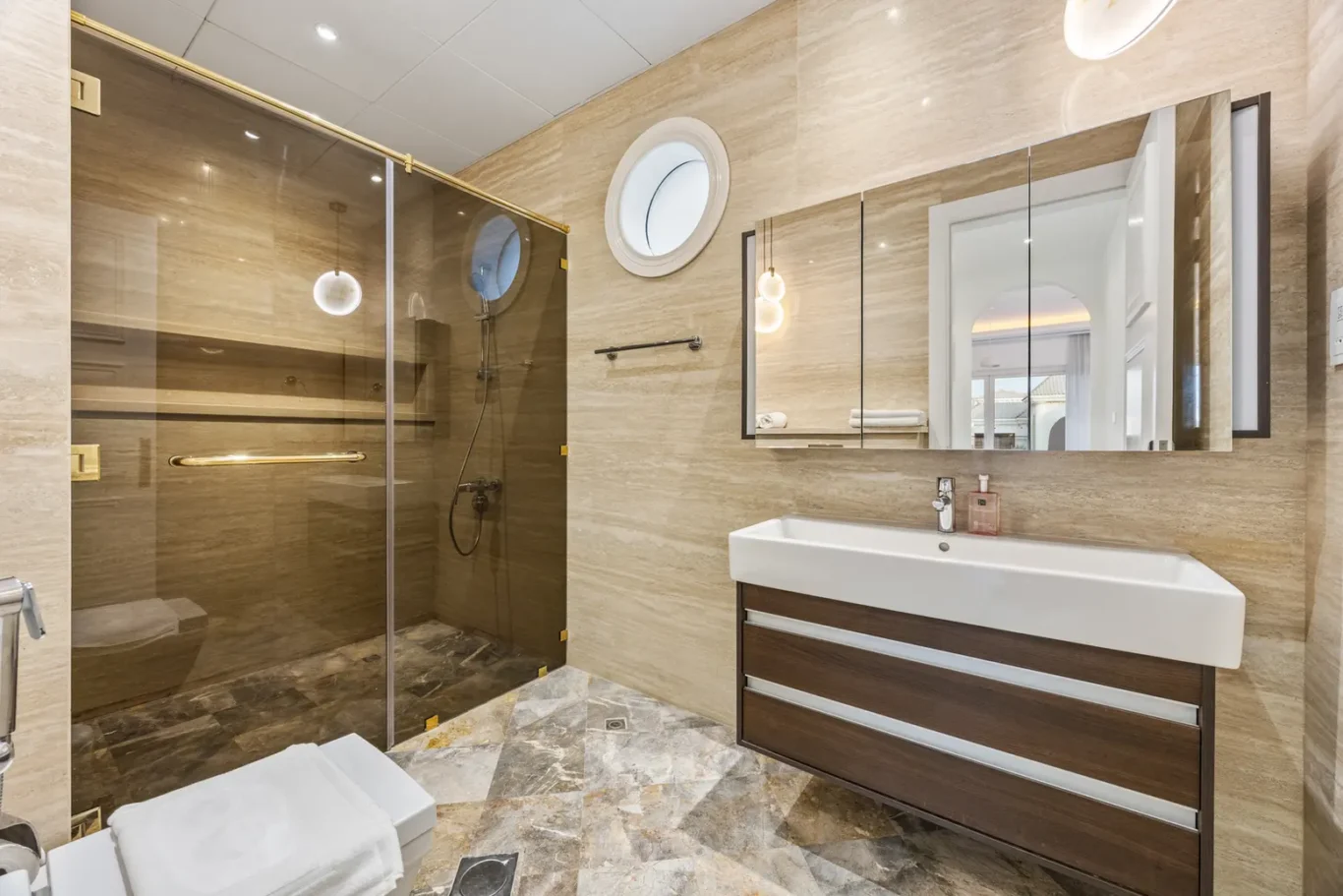 Modern beige-tiled bathroom with a glass shower enclosure, circular window, and white sink vanity with dark wood drawers.