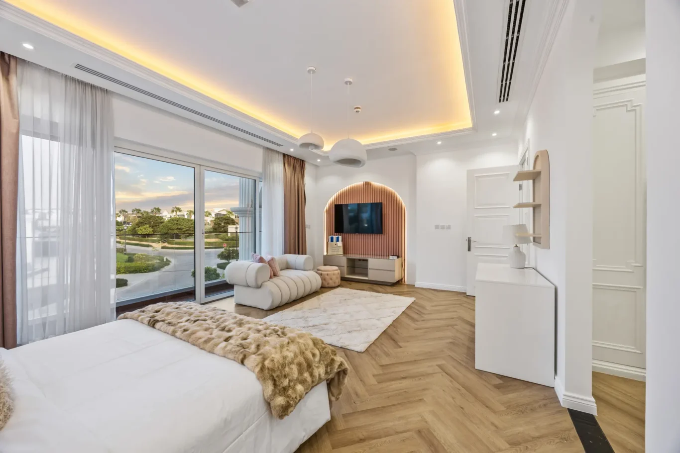 Bright modern bedroom with a large glass door to a balcony, sheer white curtains, and a white bed topped with a fur throw; a cream sofa and area rug nearby.