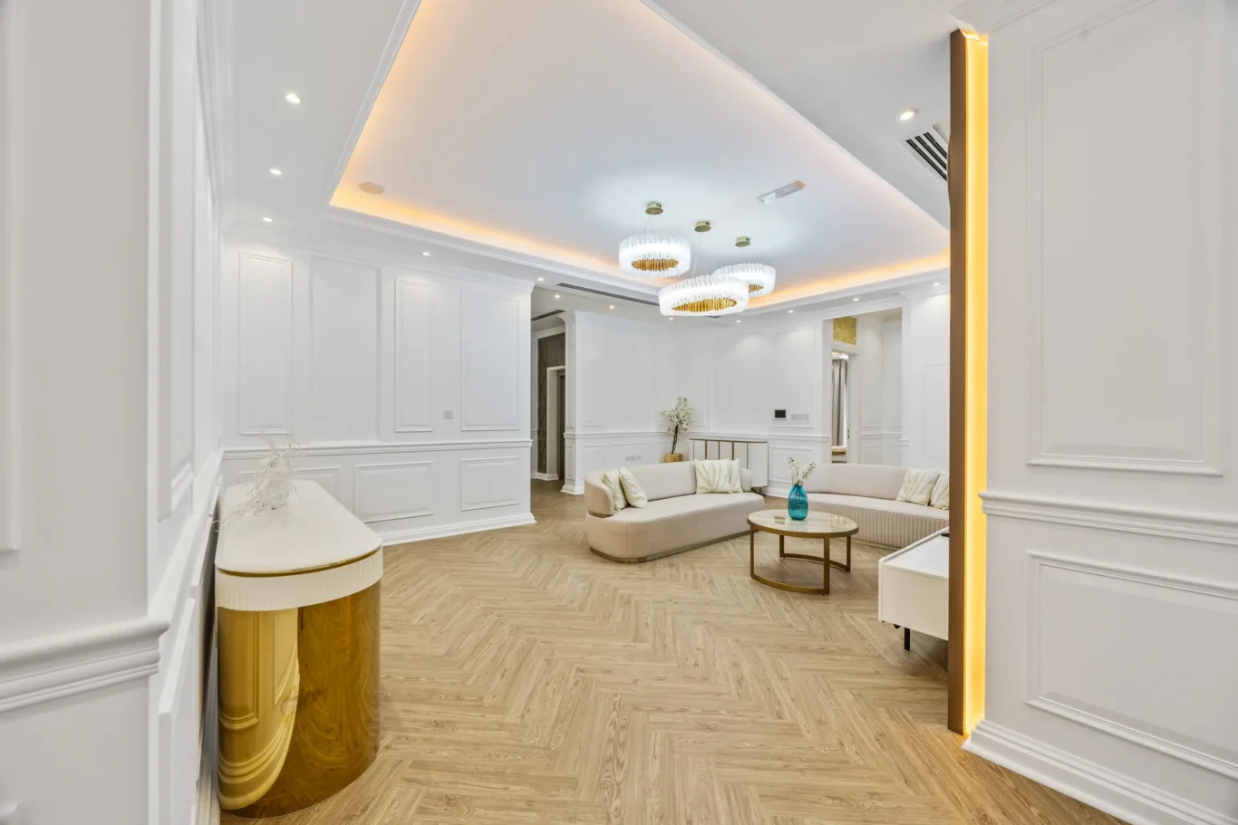 Luxurious living room with white paneled walls, herringbone wood floor, and beige curved sofas arranged around a round coffee table.