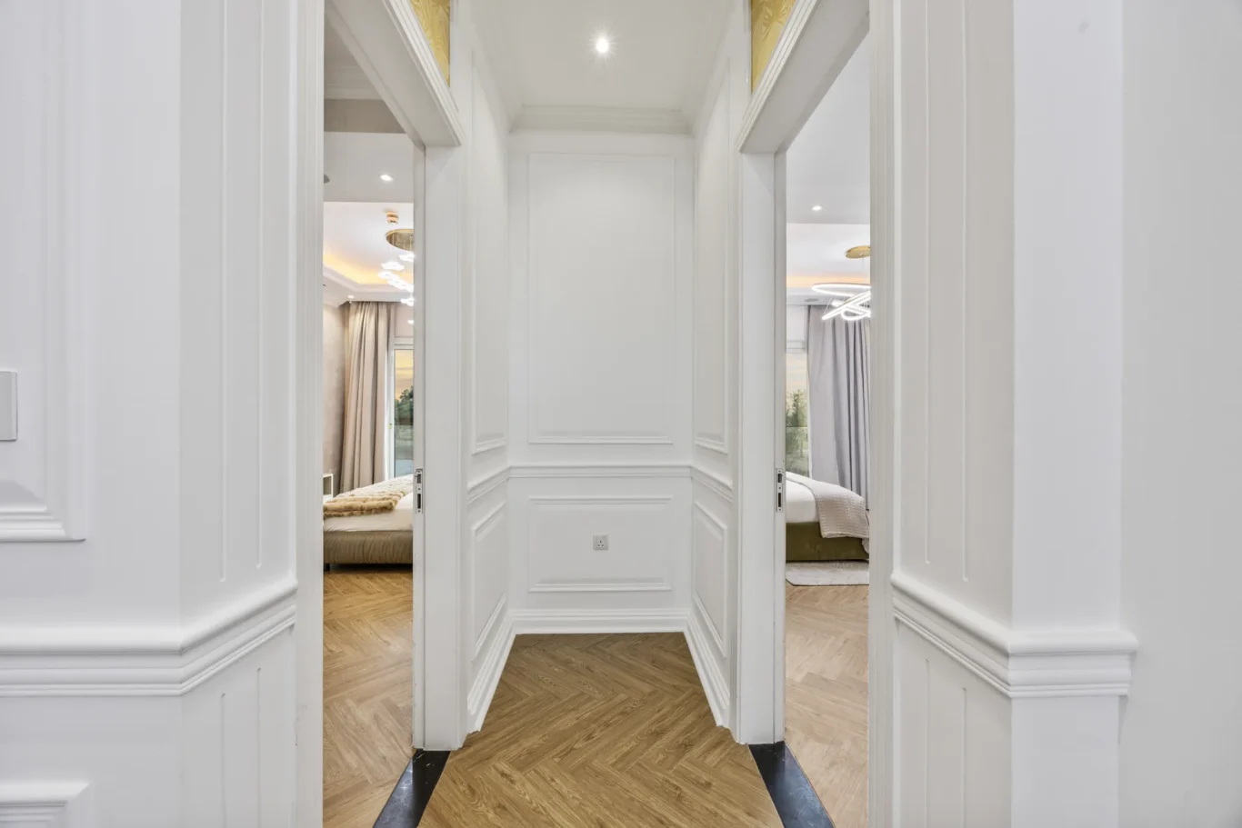 White paneled hallway leading to two bedrooms with beds visible through doorways, wood floor beneath.