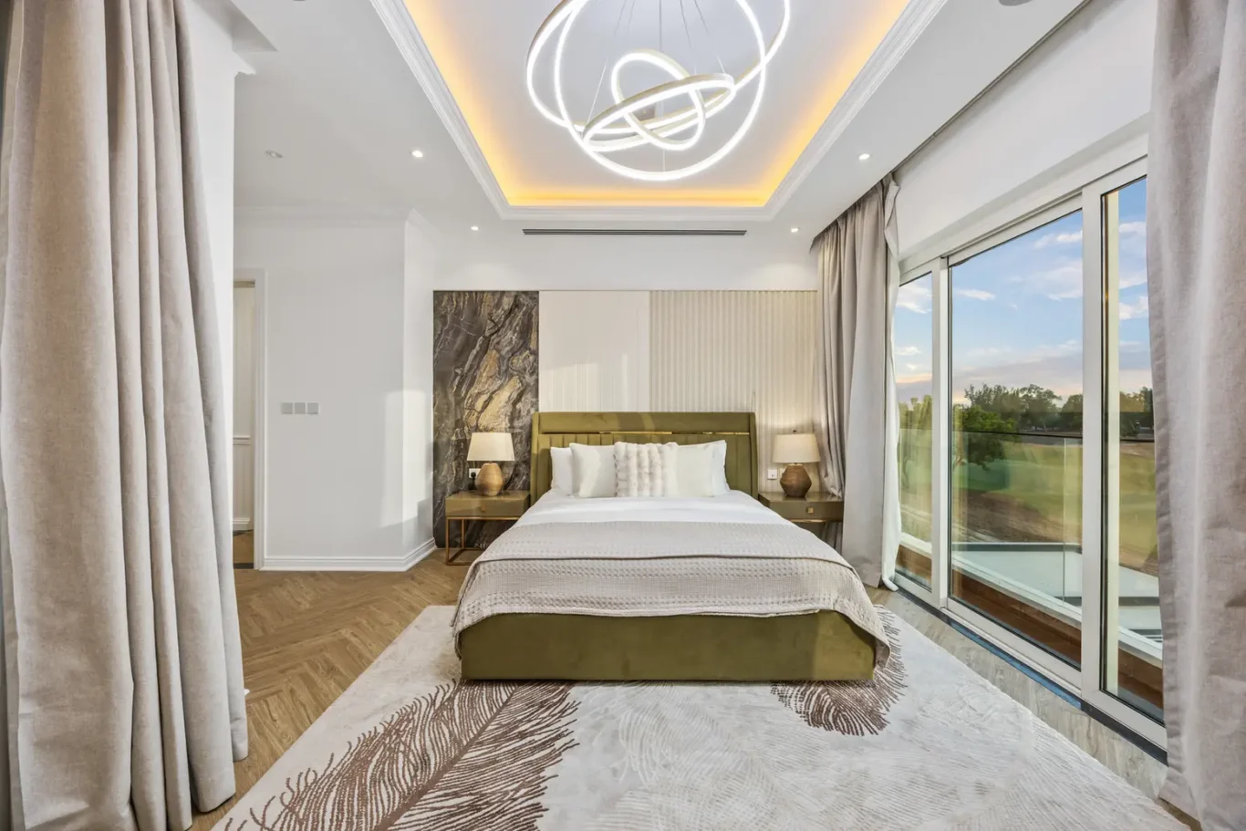 Luxurious bedroom with a king bed, olive upholstery, and matching nightstands under a modern circular chandelier in a bright suite.