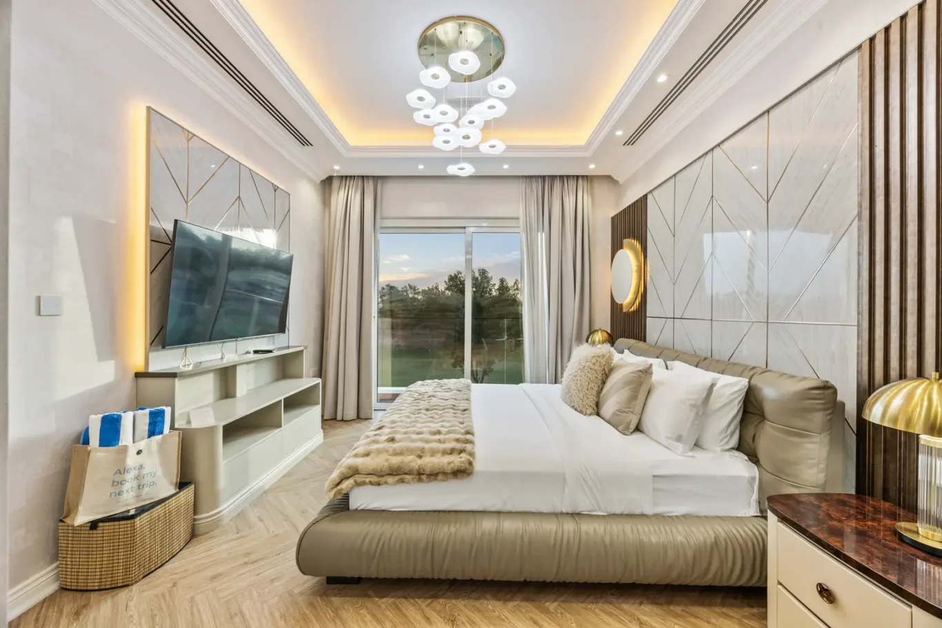 Luxurious bedroom with a low beige bed, white linens, fur throw, and a wall-mounted TV opposite a large window.