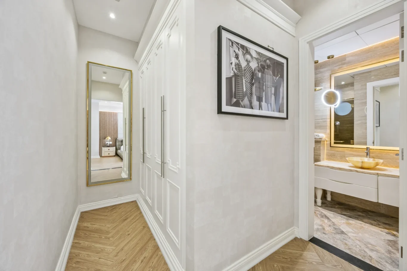 Bright hallway with built-in white wardrobes, a gold-framed full-length mirror, and a black-and-white framed art print on the wall; leads to a modern bathroom with circular mirror light and marble surfaces.