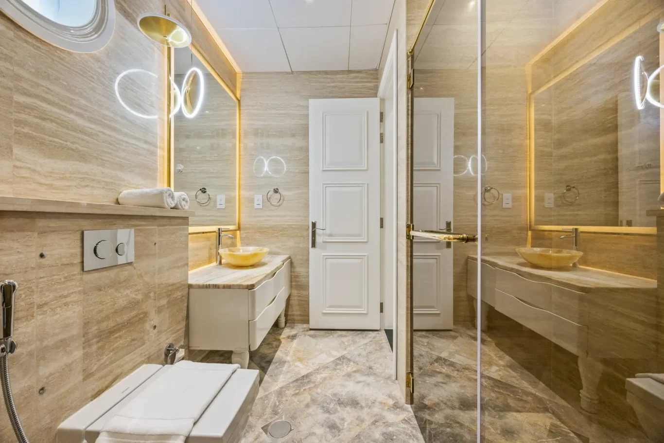 Luxurious beige marble bathroom with a double vanity, vessel sink, backlit mirrors, and a glass shower enclosure at the end of the room.