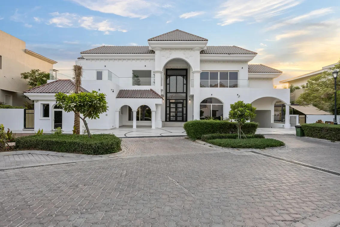 White two-story villa with arched central entrance, columns, and tiled roof, flanked by manicured hedges and a paved courtyard.