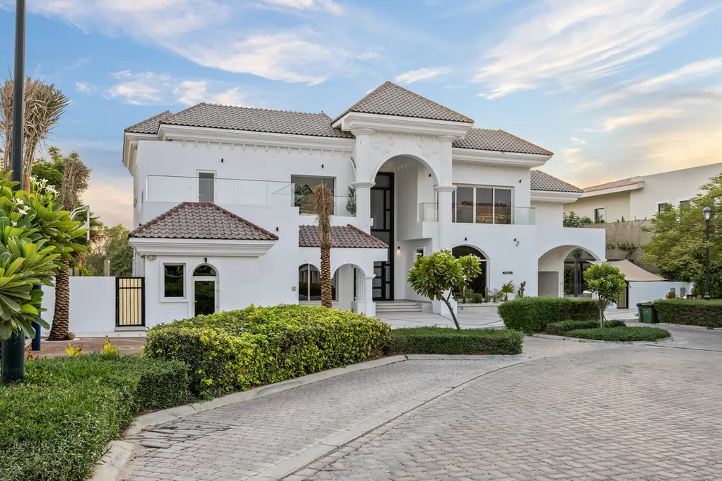 White two-story villa with arched central entrance, tiled roof, and manicured hedges along a cobblestone driveway.