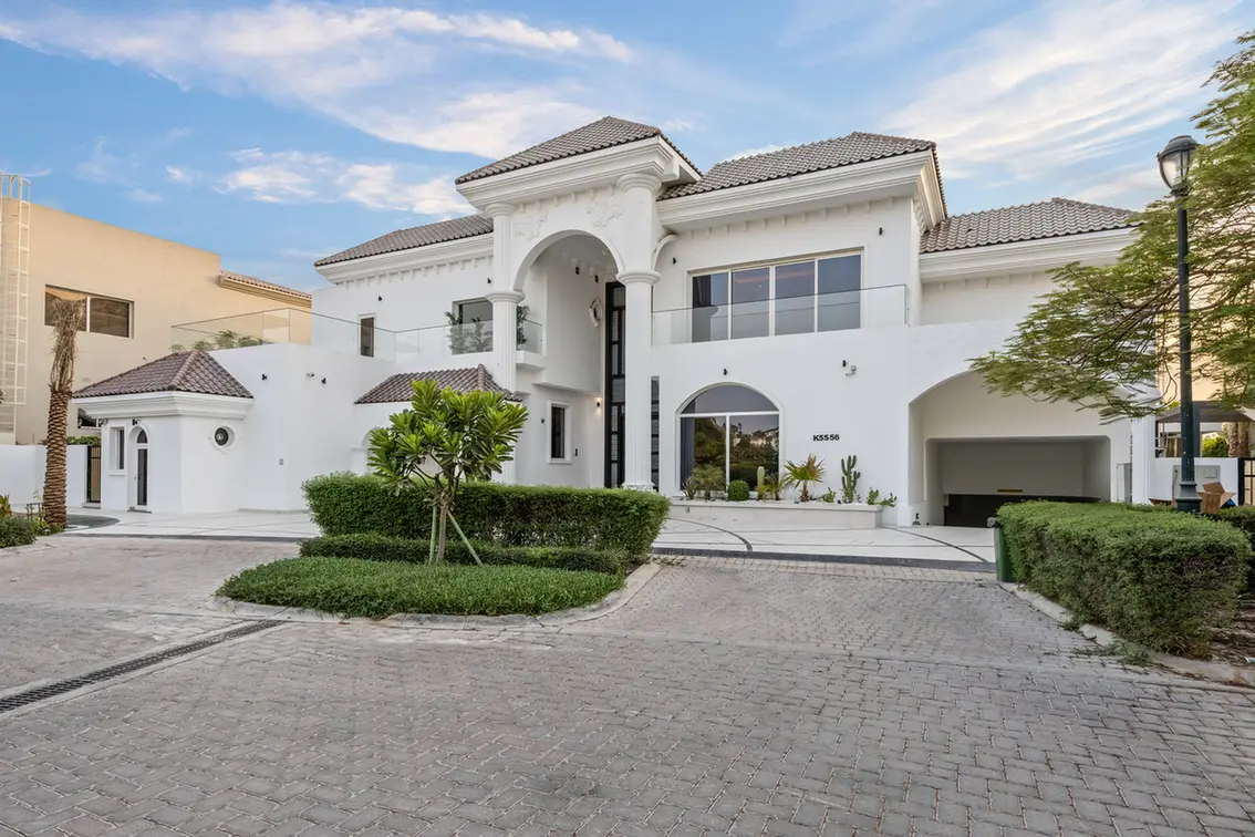 White two-story Mediterranean-style villa with an arched entryway, trimmed hedges, and a cobblestone driveway.