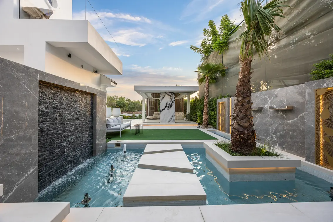 Modern backyard with a rectangular pool, white stepping stones, and palm trees beside marble walls.