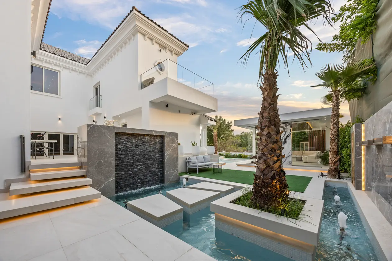 Modern white villa with a water feature, stepping-stone walkway, and palm trees in a landscaped backyard.