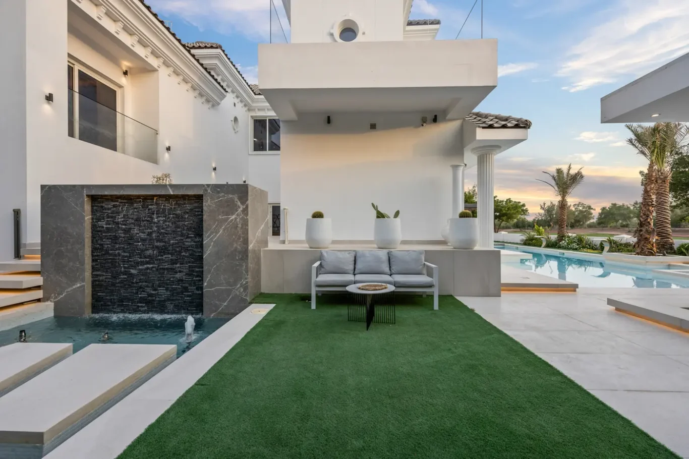 Modern white villa with an outdoor seating area by a reflecting pool and a dark stone waterfall feature.